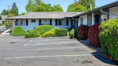 A paved parking lot in front of a white building with black trim, surrounded by a variety of trimmed bushes and hedges