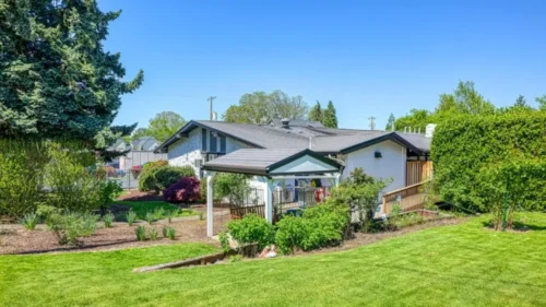 The side of a white brick building with a covered patio area and a wooden ramp, surrounded by a green lawn and trees under a clear blue sky
